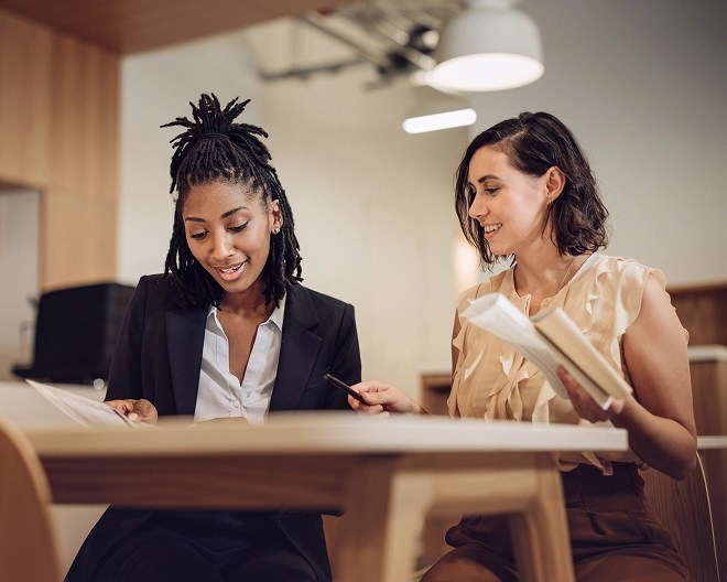 Two business professionals, one wearing a blazer over a white top and the other in a ruffled blouse with short hair, lean over a table reviewing documents together in a brightly lit, modern office setting.