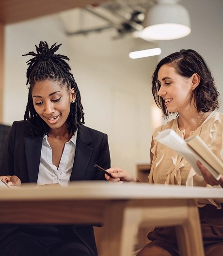 Two business professionals, one wearing a blazer over a white top and the other in a ruffled blouse with short hair, lean over a table reviewing documents together in a brightly lit, modern office setting.
