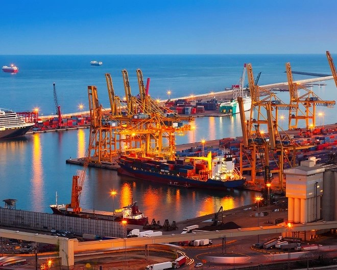 A wide shot of a shipping port lit up at dusk, with cranes, cargo containers, and docked ships visible.