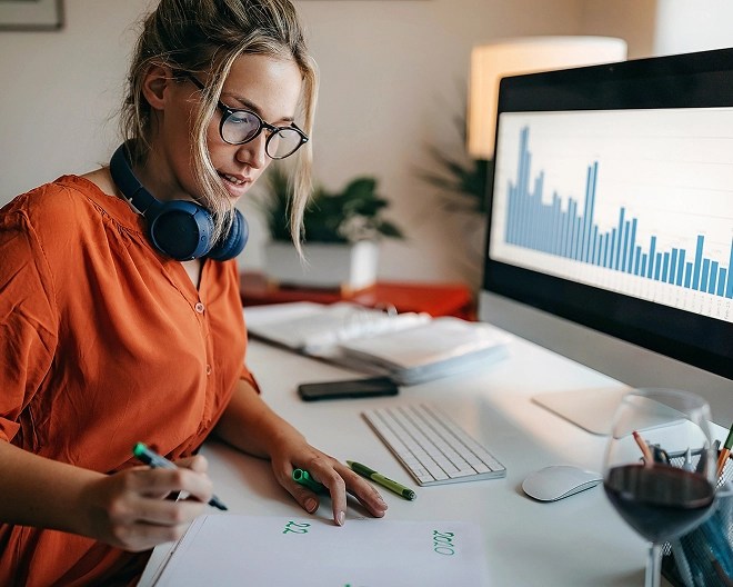 Young professional with messy updo, glasses, and headphones around her neck works from home office, jotting notes with a marker as her desktop monitor shows a blue-bar chart.