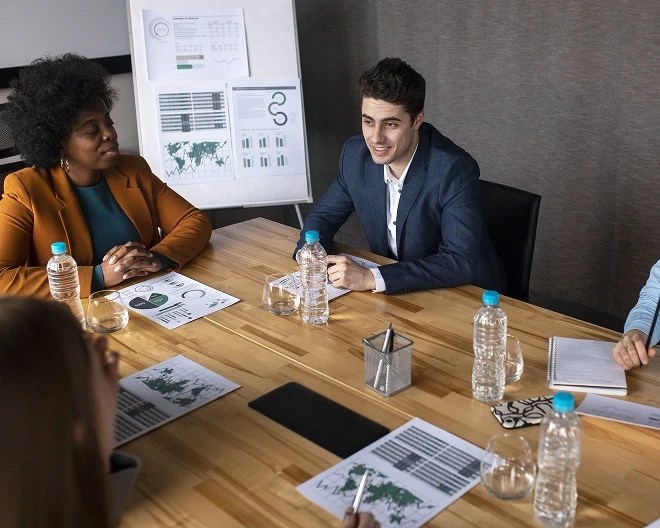 Young professionals in business casual hold a meeting around a table with water bottles and papers showing maps and charts; an easel with additional data stands in the background.
