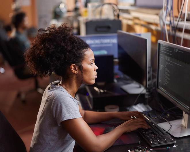 Profile of an IT professional with hair up and wearing a t-shirt, working on code in a modern office.