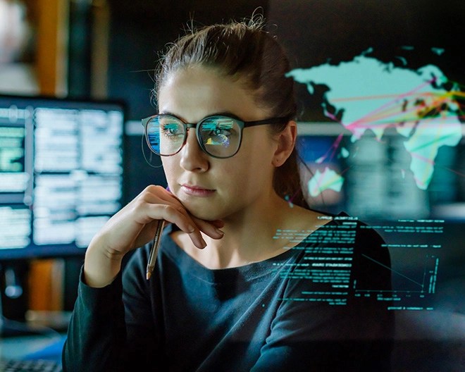 Business professional gazes at an off-screen monitor with data, coding, and map graphics reflected in her glasses.