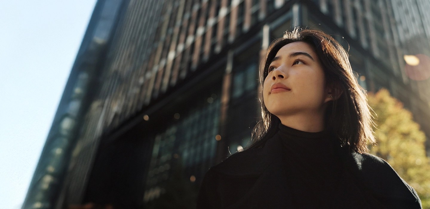 Low-angle view of a young business professional gazing upward with a hopeful expression, a tall skyscraper rising in the background against the sky.