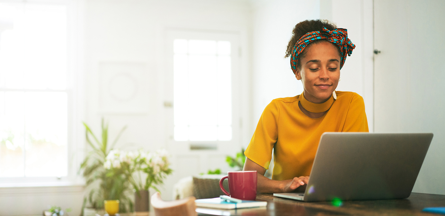 A smiling woman wearing a mustard-yellow top and patterned headband works on her laptop at a home desk, with a notebook and red mug nearby in a bright, airy room.