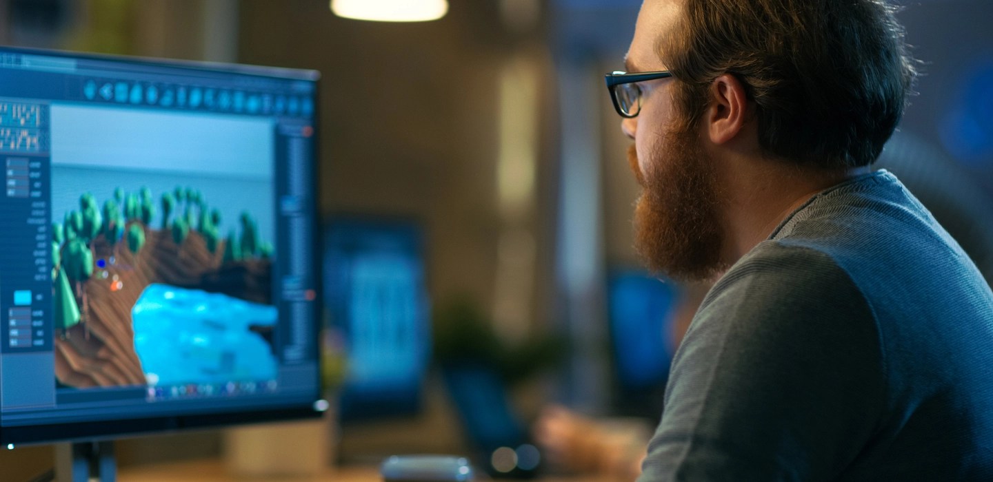 Profile of a bearded professional wearing glasses and a short-sleeved shirt, focused on his computer screen displaying 3D modelling.