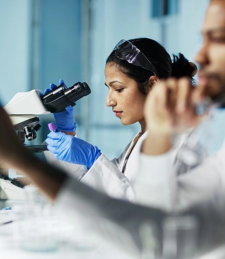 Lab researcher wearing blue latex gloves and a white coat holds a test tube while leaning forward to examine a sample through a microscope.