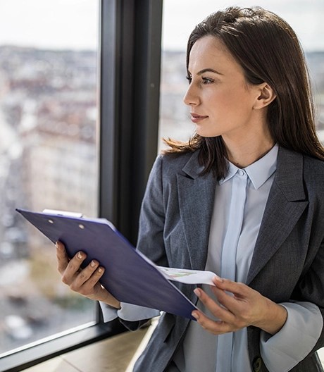Young business professional in a blazer stands on the top floor of a city office, holding a clipboard and gazing out over at the city below through tall windows.