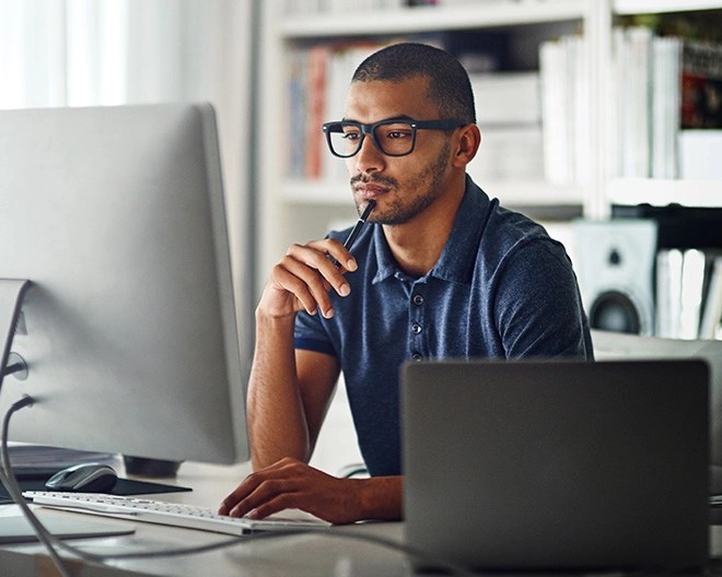 Focused online student sits in a bright home office, looking intently at his desktop computer with a pen resting on his chin.