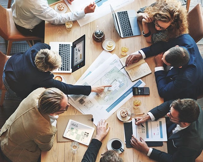 Bird’s-eye view of business professionals gathered around a table during a meeting, with laptops and notebooks open, and business plans scattered across the table that they are referencing.
