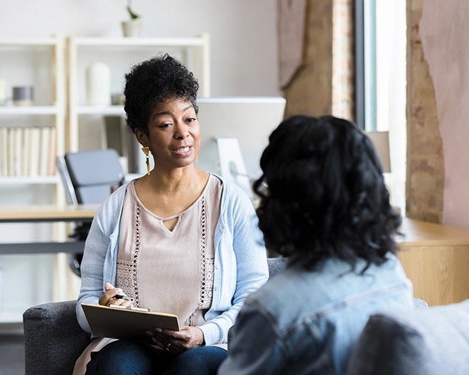 Counseling professional attentively addresses a client while taking notes on a notepad in a bright, well-lit office.