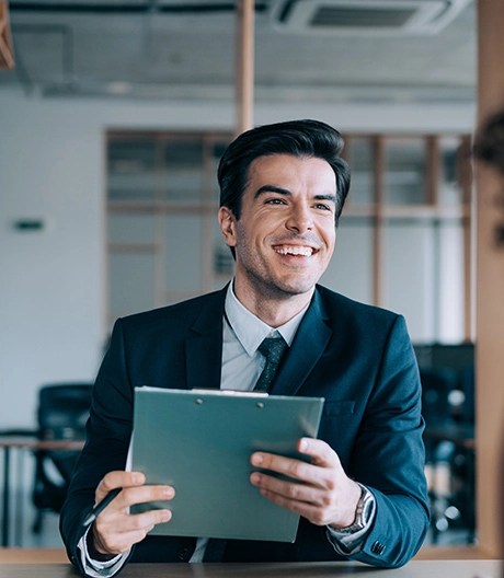 Charming business professional in a suit smiles at a colleague during a business meeting while holding a clipboard.