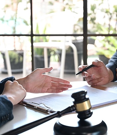 Close-up of two lawyers discussing a case in an office; one points to information on a clipboard while the other holds a pen, with a gold-ribboned gavel softly blurred in the foreground.