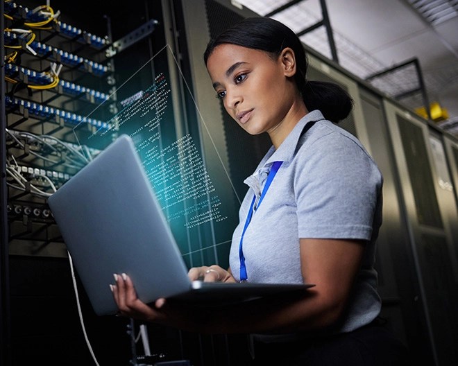 Female cybersecurity analyst reviewing code on a laptop while standing between server racks in a data centre.