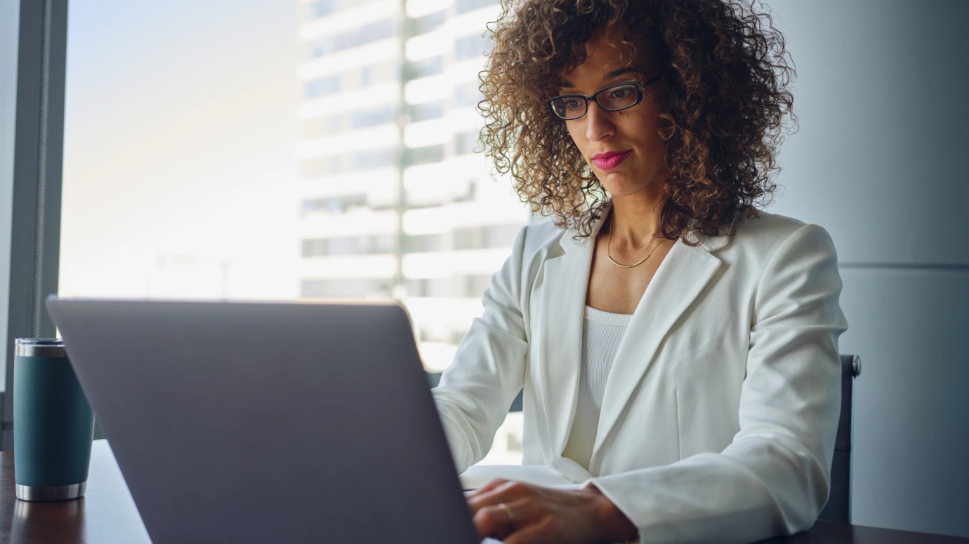 Business professional in a white suit working on her laptop in a high-rise office with large windows and city views in the background.