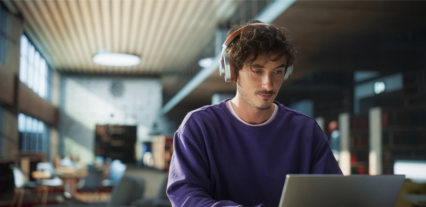 Online student wearing a purple sweatshirt and headphones, focused on his laptop while studying in a university library.
