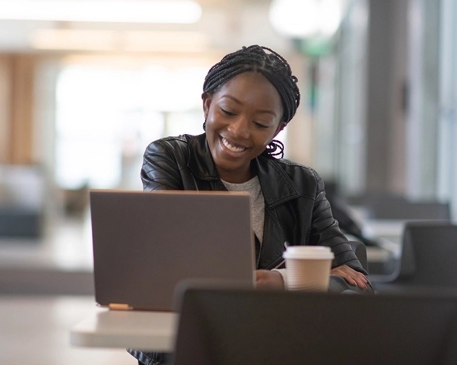 A student in a leather jacket smiling at her laptop, with a coffee on the desk.
