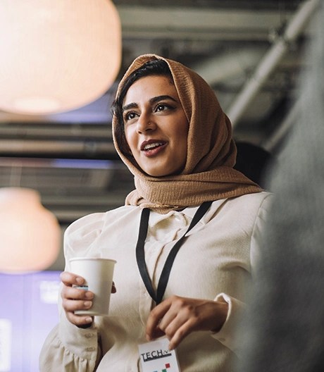A woman in a hijab holding a coffee, speaking to two people under a large light.
