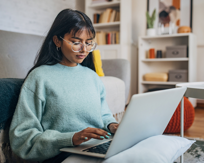 A student sitting on a sofa in a blue sweater, typing on her laptop with a pillow on her lap.