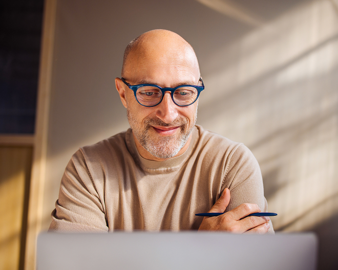 A bald man with glasses looking at a laptop, holding a pen.