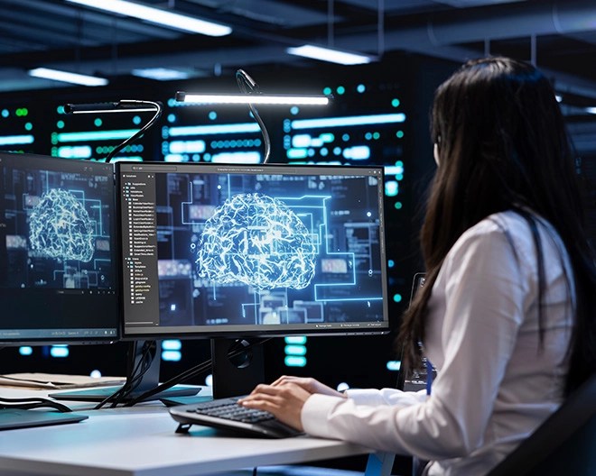 A woman sitting at a desk looking at two monitors displaying bright technology.