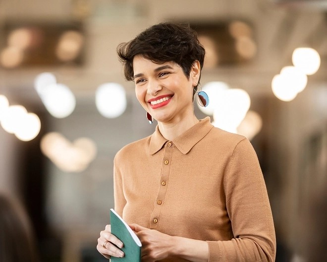 A woman in a brown dress holding a green notebook, smiling.