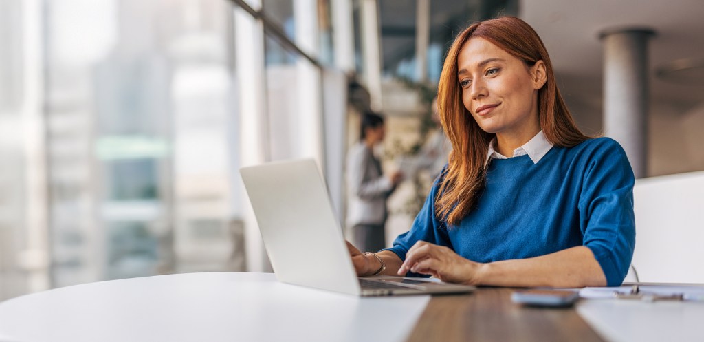 A woman in a blue sweater sitting at a desk, typing on a laptop.
