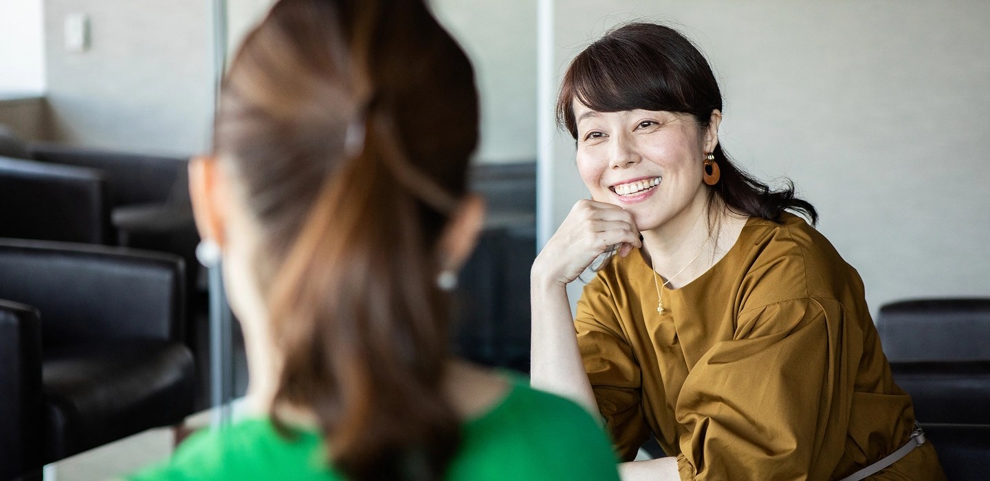 A woman in a yellow shirt smiling while talking to another woman wearing a green shirt.