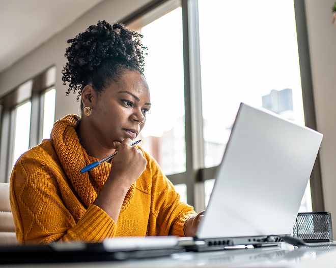 A woman in a yellow sweater holding a pen and looking at her laptop.