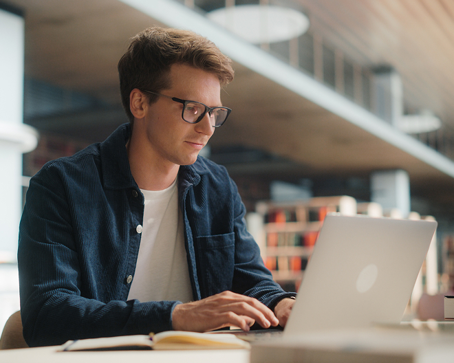 A student wearing glasses typing on his laptop at a library desk, with a notebook nearby.