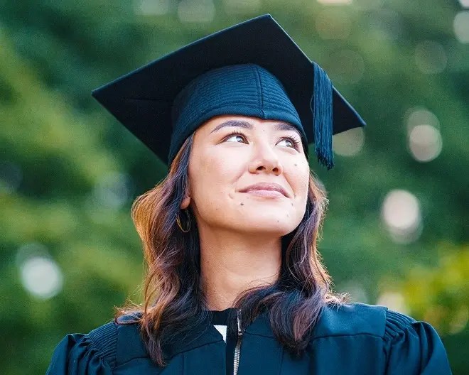 Graduate student wearing a navy cap and gown, smiling softly and looking up hopefully.