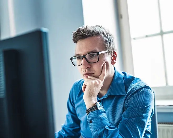 Young professional wearing a blue button-up and glasses, rests his hand on his chin as he works intently on the computer.