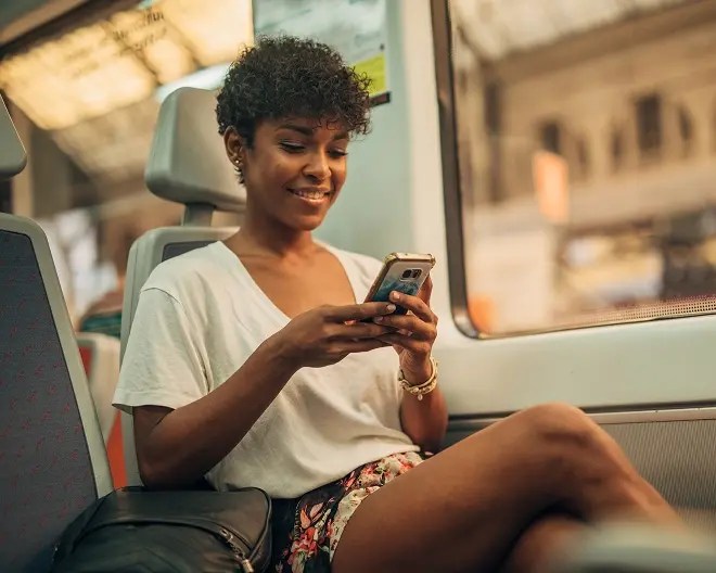 Person wearing casual white t-shirt and holding phone, smiles as she looks at her device while on a commuter train.