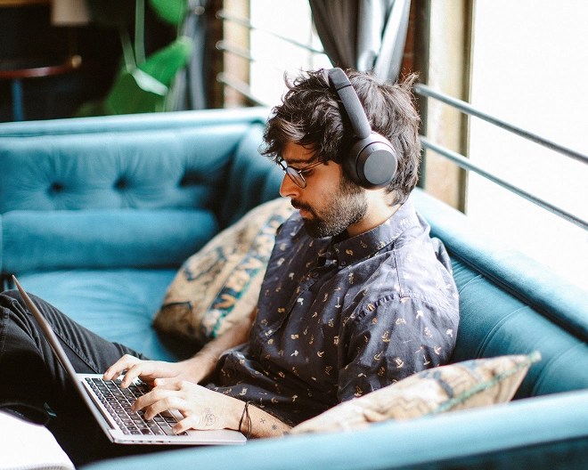 Man with a beard, dressed in casual clothes, wearing glasses and headphones, working from home on his laptop while sitting on his couch.