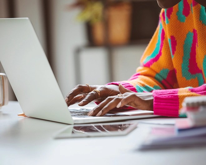 Close-up of a woman wearing a colourful pink, teal, and orange sweater, working on her computer resting on a white tabletop.
