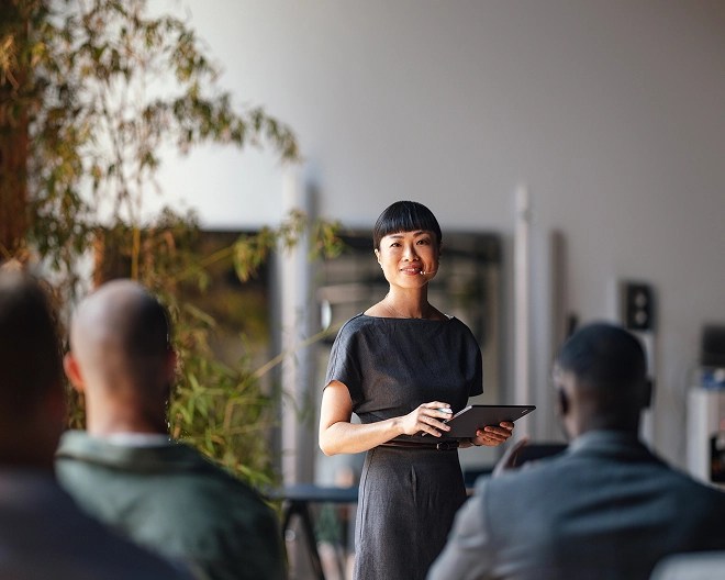 Smiling businesswoman with bangs, dressed in business casual, presenting during a meeting while her colleagues watch from across the table.