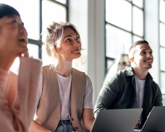 Young business professionals with laptops smile as they watch an off-camera presenter during a meeting.