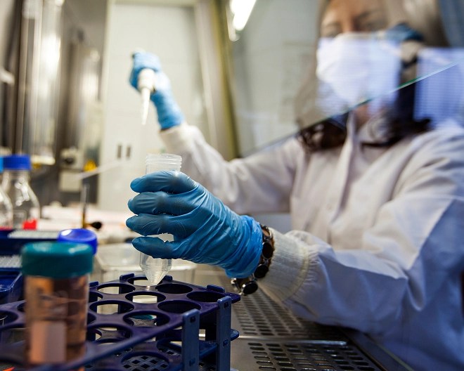 lab technician wearing a white coat and blue gloves uses a pipette to transfer liquid into a conical tube at a laboratory workbench filled with scientific equipment.