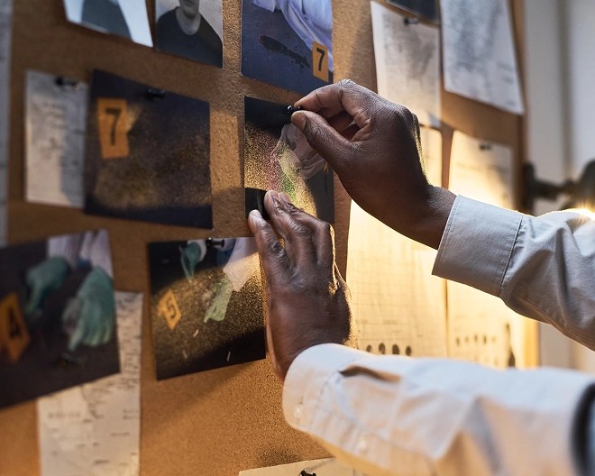 Close-up of hands pinning a crime scene photo to a bulletin board, suggesting investigative work in progress.