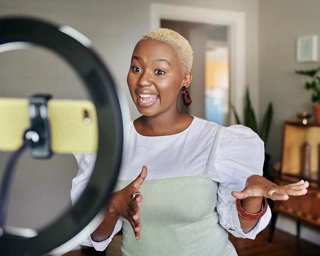 A content creator stands in front of a ring light and smartphone on a tripod, recording or streaming video in a stylish room decorated with plants, framed art, and shelving.