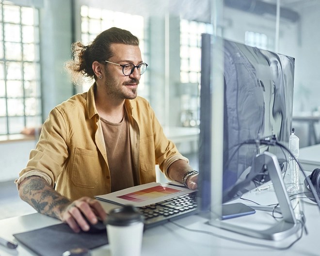 Person in a yellow shirt with visible tattoos works intently at a desk with a large computer monitor, surrounded by office equipment in a modern workspace with natural light.