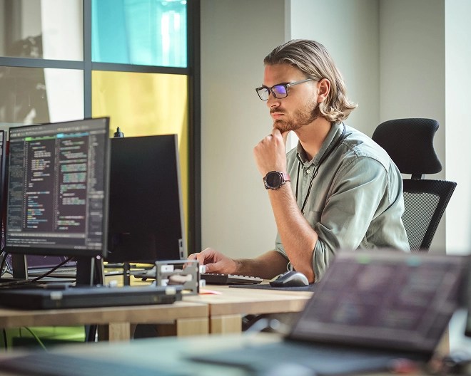 Person in casual clothes and glasses sits in brightly lit tech office space as he codes on his computer.