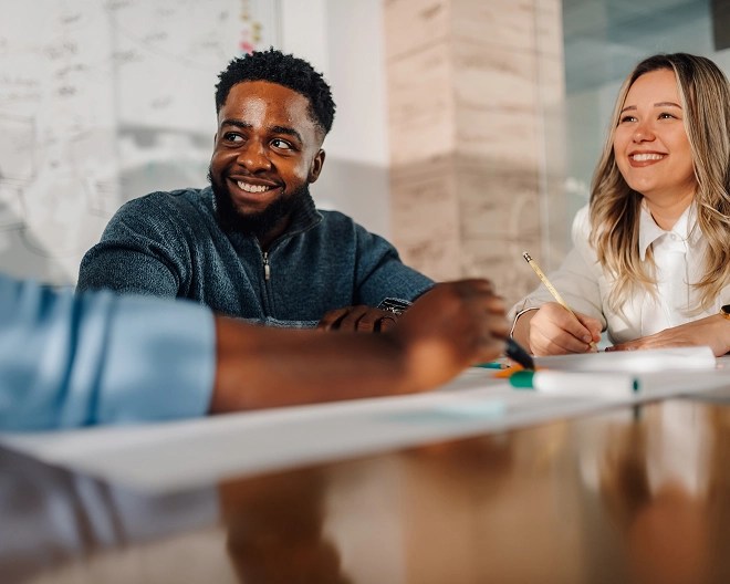 Person in a knit half-zip sits at a meeting table and smiles to his colleagues out of frame