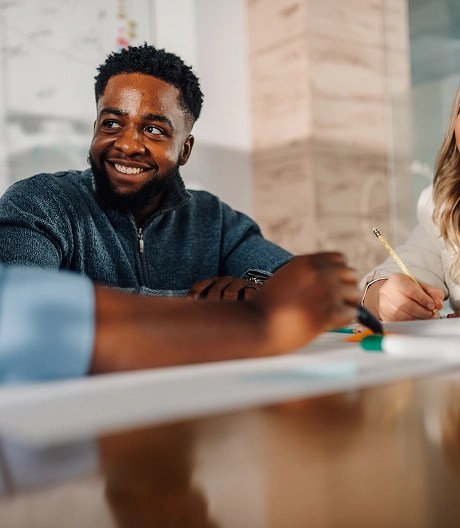 Person in a knit half-zip sits at a meeting table and smiles to his colleagues out of frame