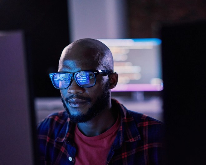 Close-up of a person coding in a dimly lit office space, his work reflected in his glasses and a computer with code in the background.