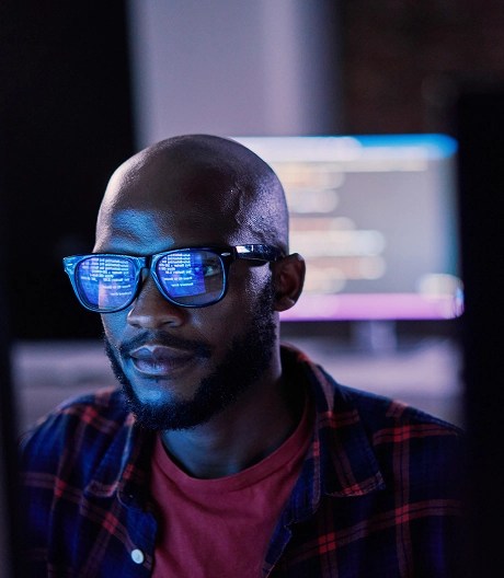 Close-up of a person coding in a dimly lit office space, his work reflected in his glasses and a computer with code in the background.