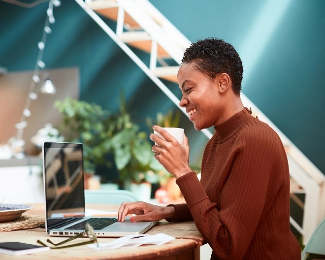 Smiling woman in a brown sweater sitting at a round table, holding a white mug and working on a laptop in a bright, modern home interior with plants and a staircase in the background.