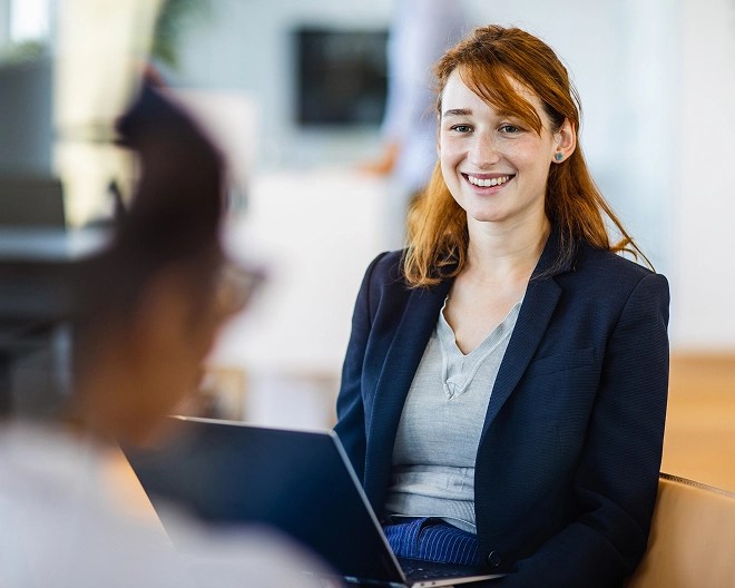 Young woman in a navy blazer smiling during a conversation, seated with a laptop on her lap in a modern office setting, with another person blurred in the foreground.