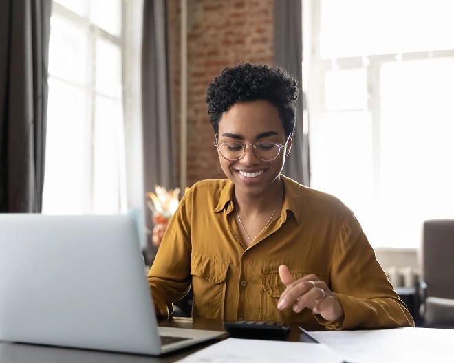 Smiling woman in a light brown blouse, punching numbers on a calculator next to her open laptop.
