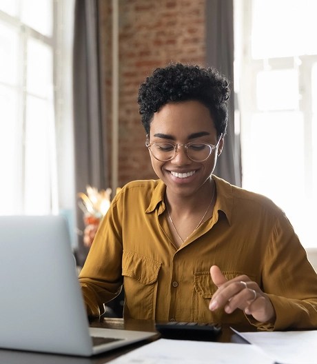 Smiling woman in a light brown blouse, punching numbers on a calculator next to her open laptop.